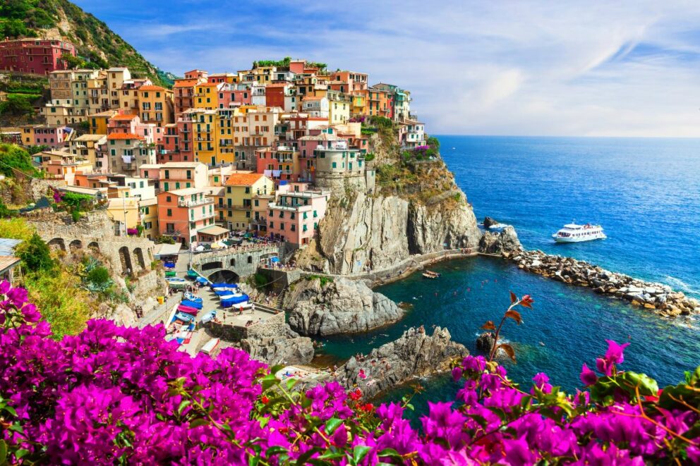 Vibrant cliffside village in Cinque Terre, Italy, with colorful houses stacked above a turquoise cove and bright pink flowers in the foreground overlooking the Mediterranean Sea.