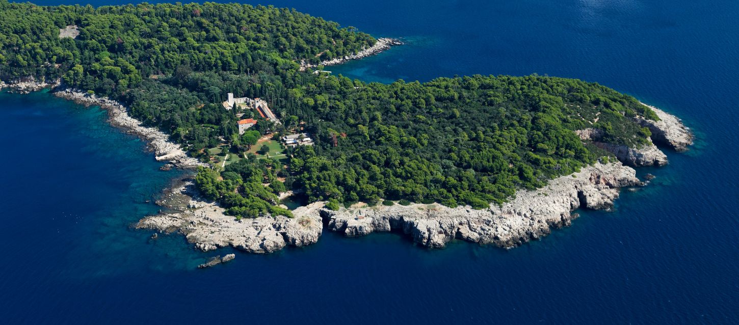 Aerial view of Lokrum Island, in the Adriatic Sea. Rocky coastline and dense green vegetation, with a few buildings, possibly a monastery, visible on the shore. The surrounding sea is a deep blue color.