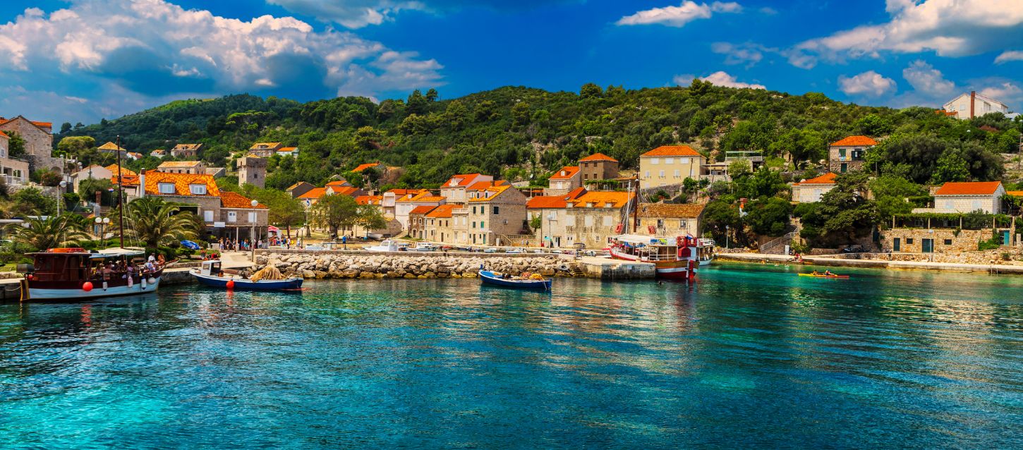 picturesque coastal village with houses featuring red-tiled roofs, nestled on a green, hilly shoreline. The foreground is dominated by clear turquoise water where several boats are moored near a rocky harbor wall. The sky is bright blue with some white clouds, suggesting a sunny day.
