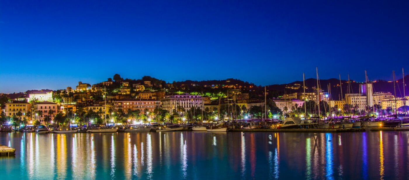 Night view of La Spezia, Italy, with illuminated waterfront buildings and sailboats reflected in the calm harbor waters under a deep blue sky.