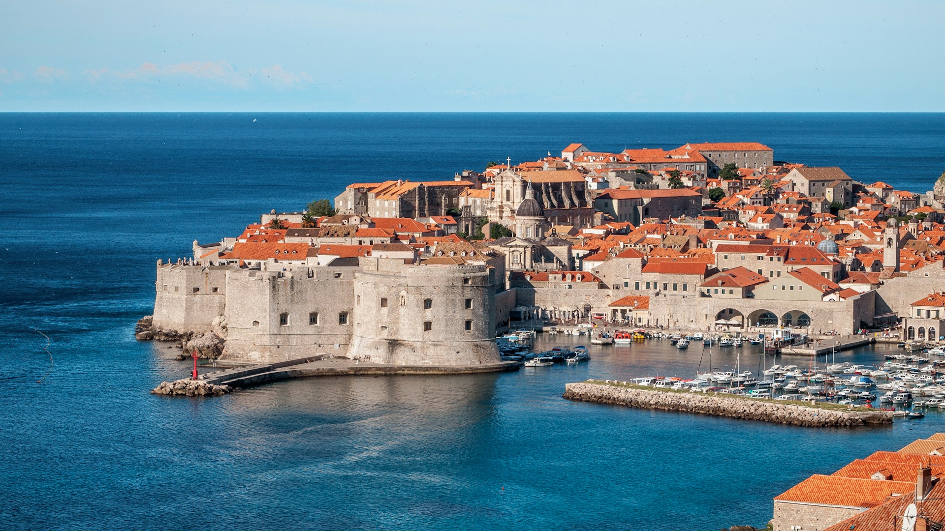 Aerial view of Dubrovnik, showing the walled city with its orange roofs, the medieval walls surrounding the old town, and part of the harbor with boats moored on the turquoise waters of the Adriatic Sea.