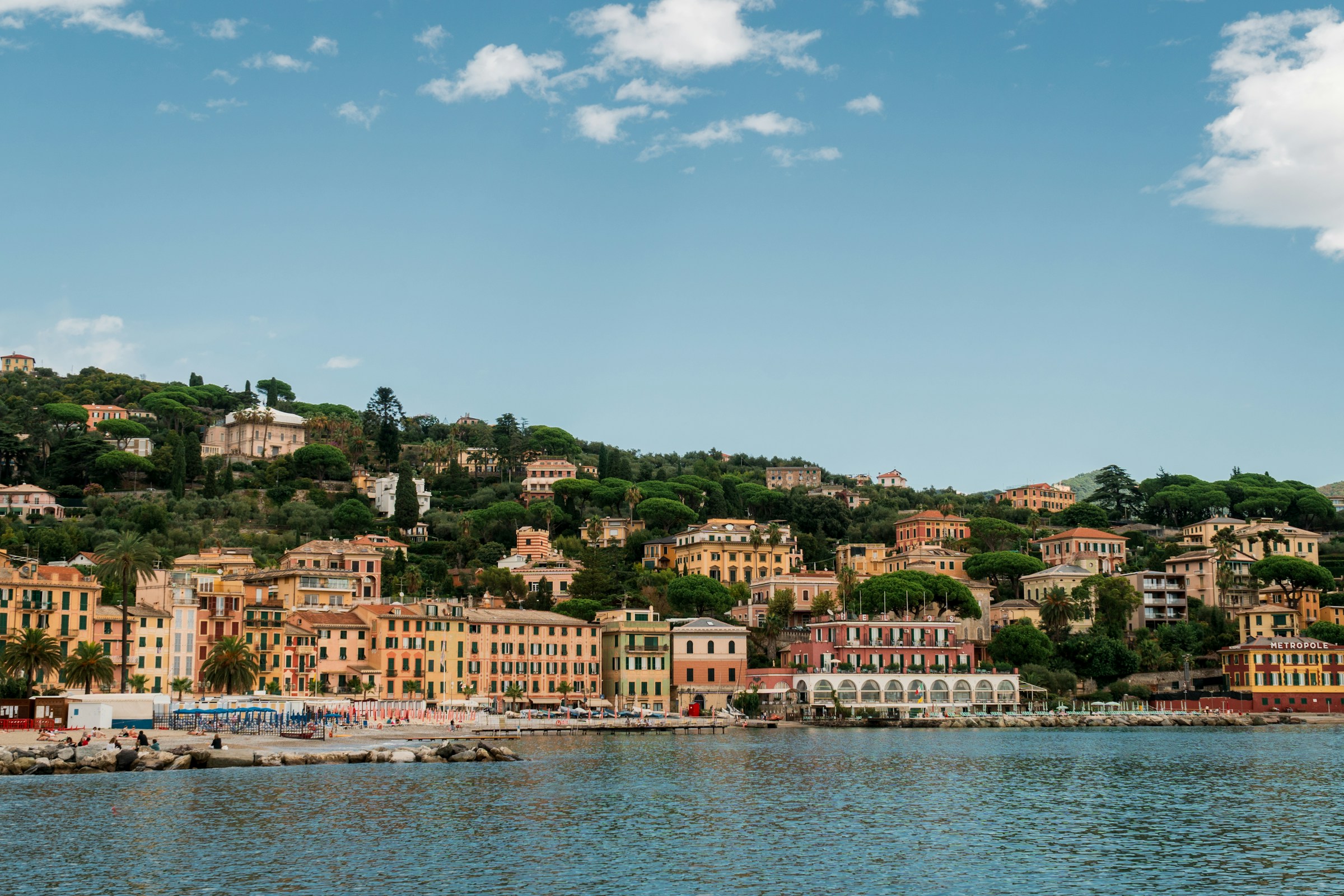 Colorful hillside buildings along the Italian Riviera overlooking calm blue waters, with lush greenery and palm trees under a partly cloudy sky.